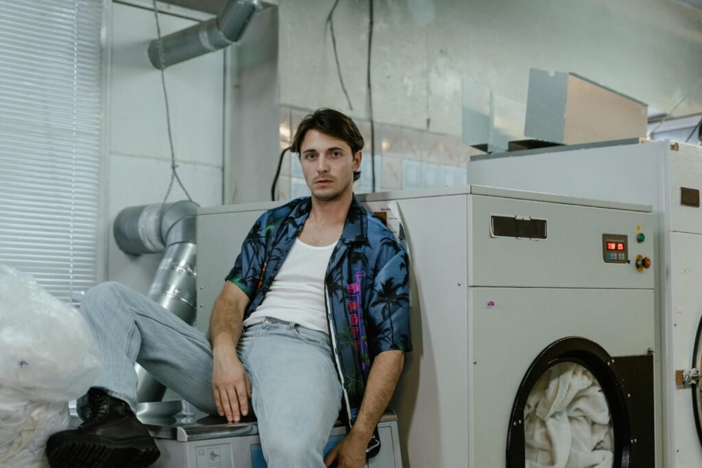 Young man sitting casually on a dryer in an industrial laundromat setting.